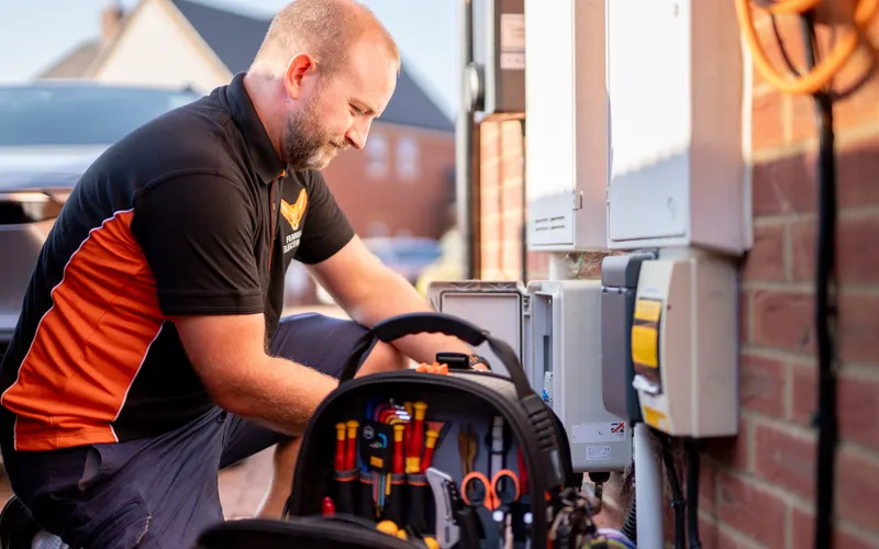 The electrician kneeling at his tool bag beside a consumer unit on a brick wall